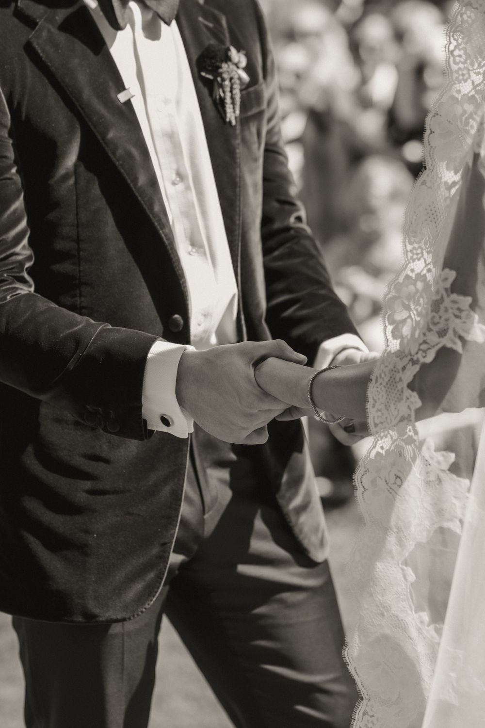 Black and white photo of a groom in a suit holding hands with a bride in a lace wedding dress during ceremony.