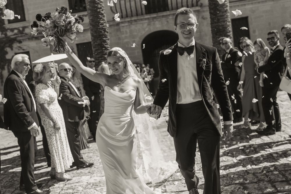 Black and white photo of a joyful bride and groom walking through confetti after their outdoor wedding ceremony.