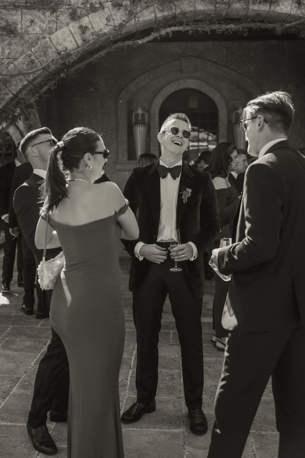 Black and white photo of elegantly dressed guests socializing outdoors at a formal event, man in tuxedo with sunglasses smiling.