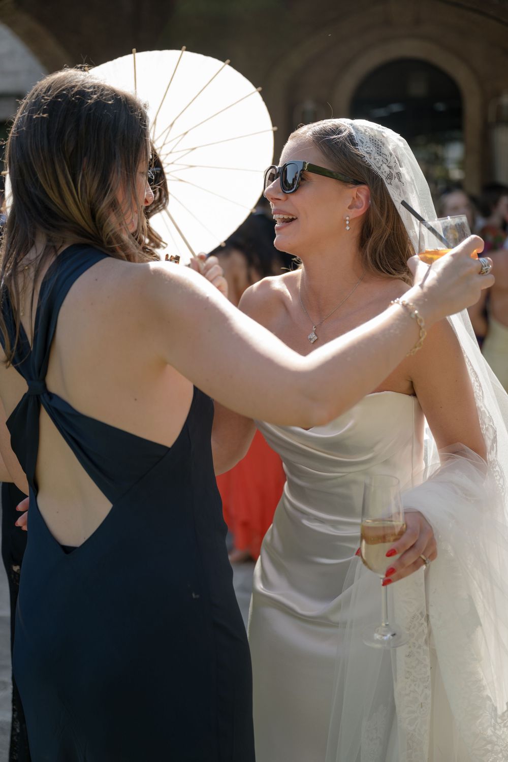 Bride in white gown and veil with sunglasses toasts champagne with guest in navy dress at outdoor wedding.