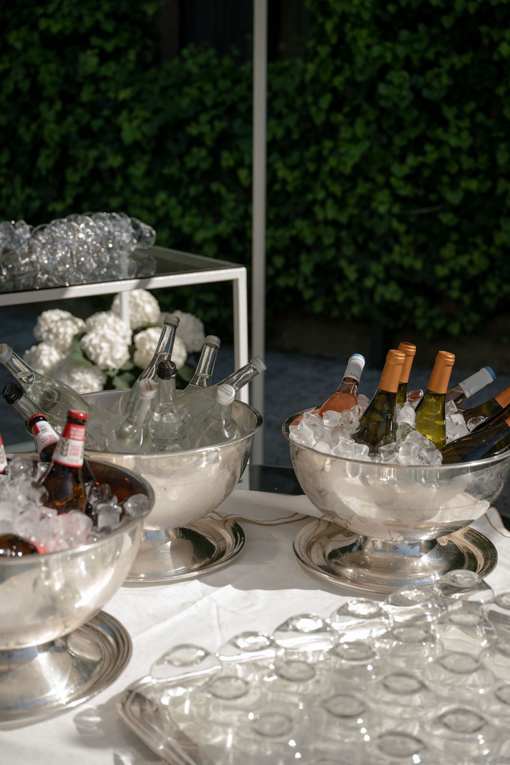 Silver ice buckets filled with wine and beer bottles on a white tablecloth at an elegant outdoor event.