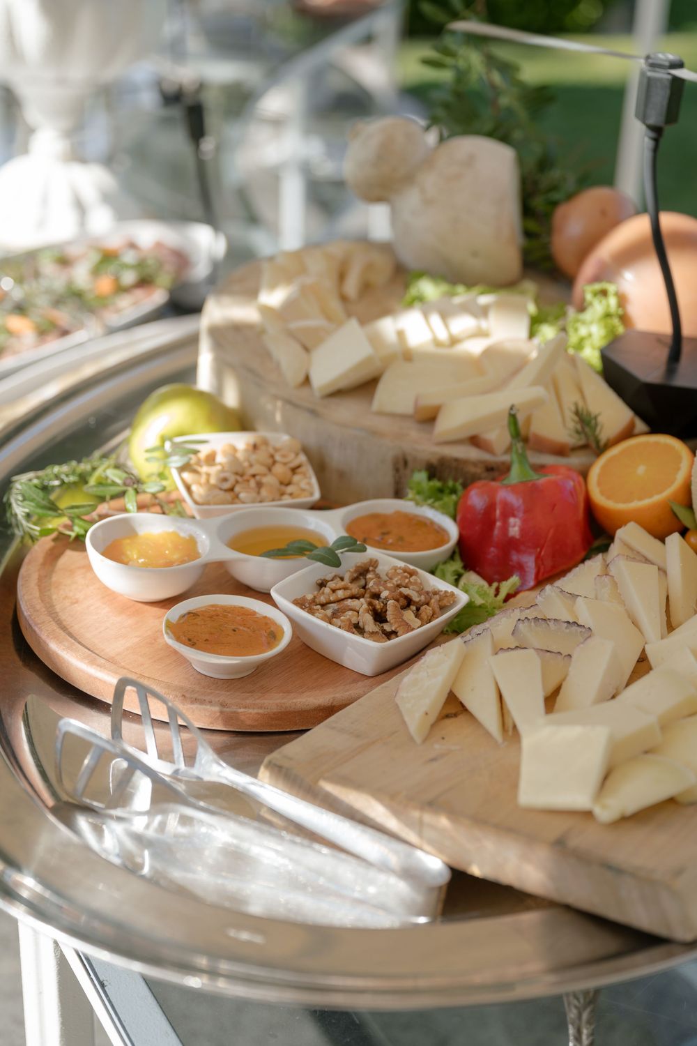 Elegant cheese board with assorted cheeses, nuts, honey dips, fresh fruits, and herbs on wooden boards at an outdoor event.