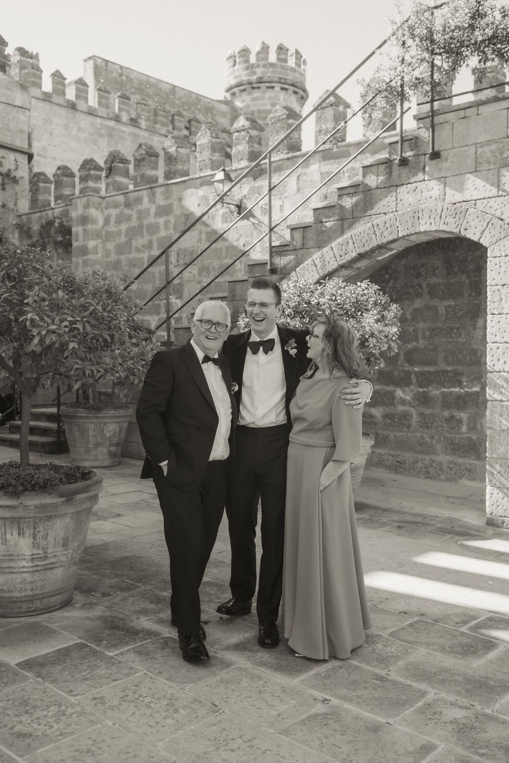 Black and white photo of three elegantly dressed people posing in front of a stone castle courtyard.