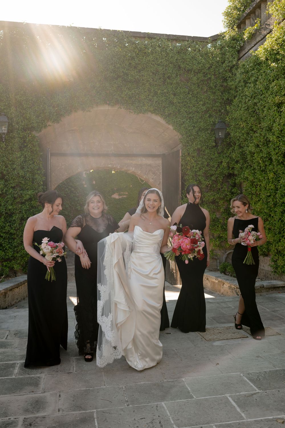 Bride in white gown walks with bridesmaids in black dresses, holding bouquets, in front of ivy-covered stone archway.