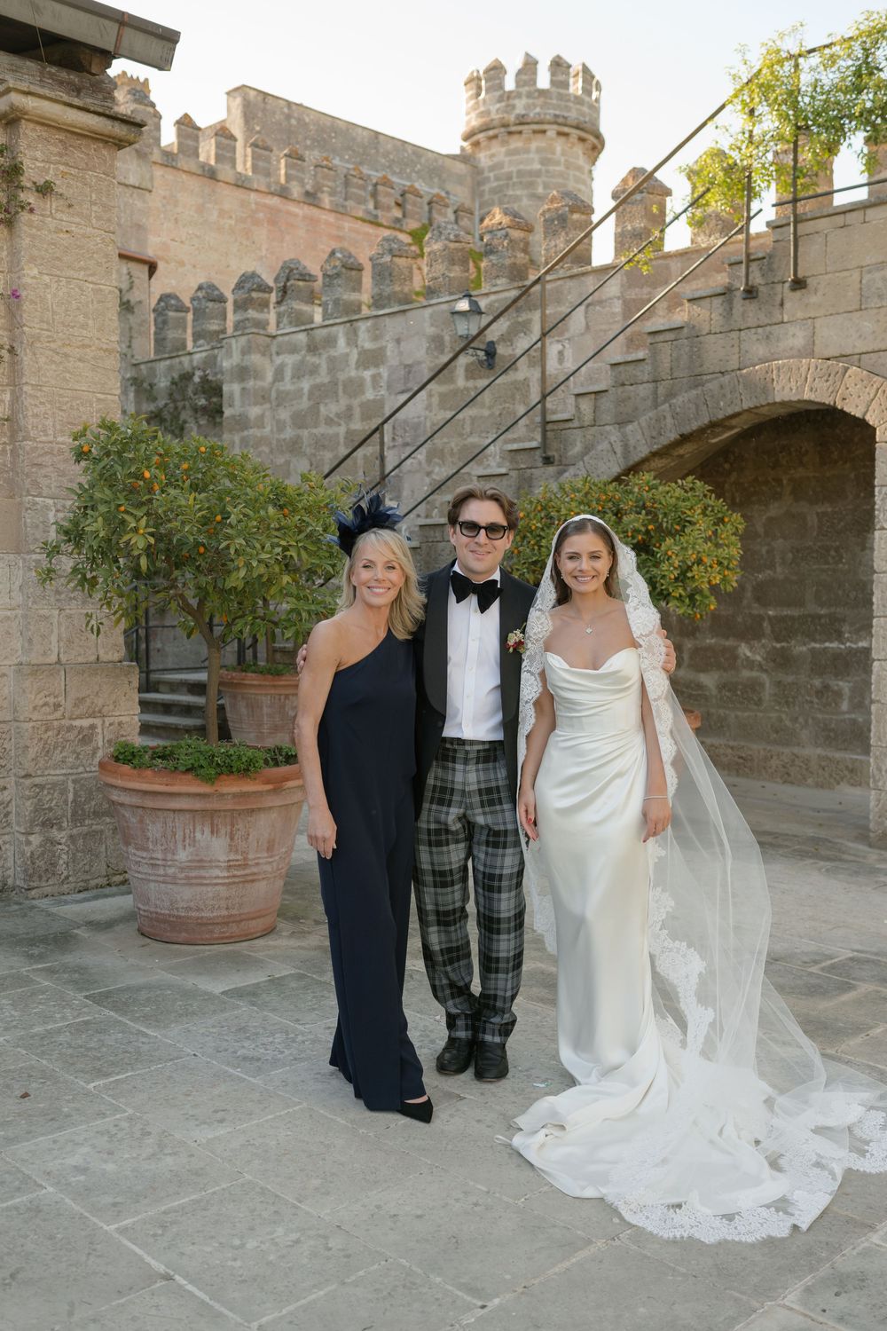 Wedding party poses before a castle, bride in white gown with veil, groom in plaid pants, woman in navy dress.