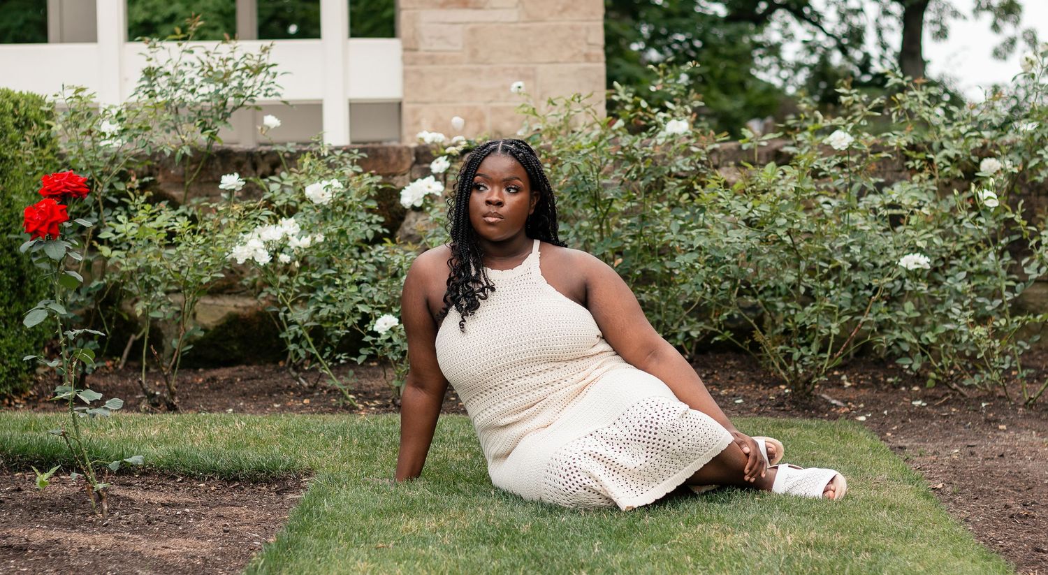 Woman in cream dress sitting gracefully on green grass surrounded by blooming white and red roses in a garden.