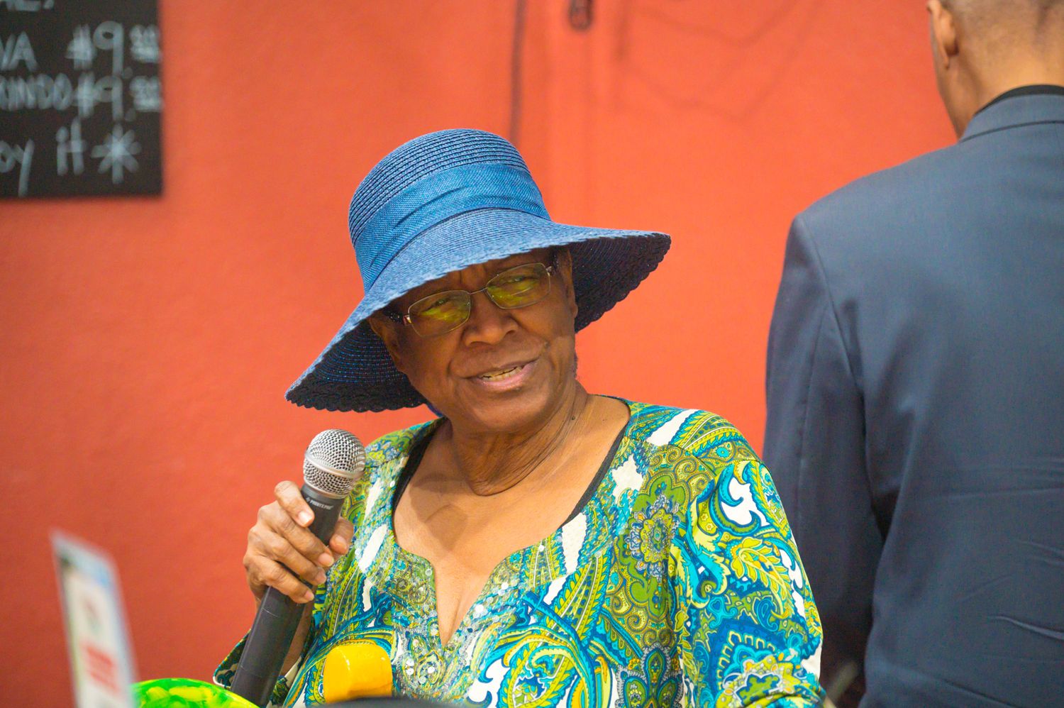 Elderly woman wearing a blue sun hat speaks into a microphone outdoors in a colorful floral top.