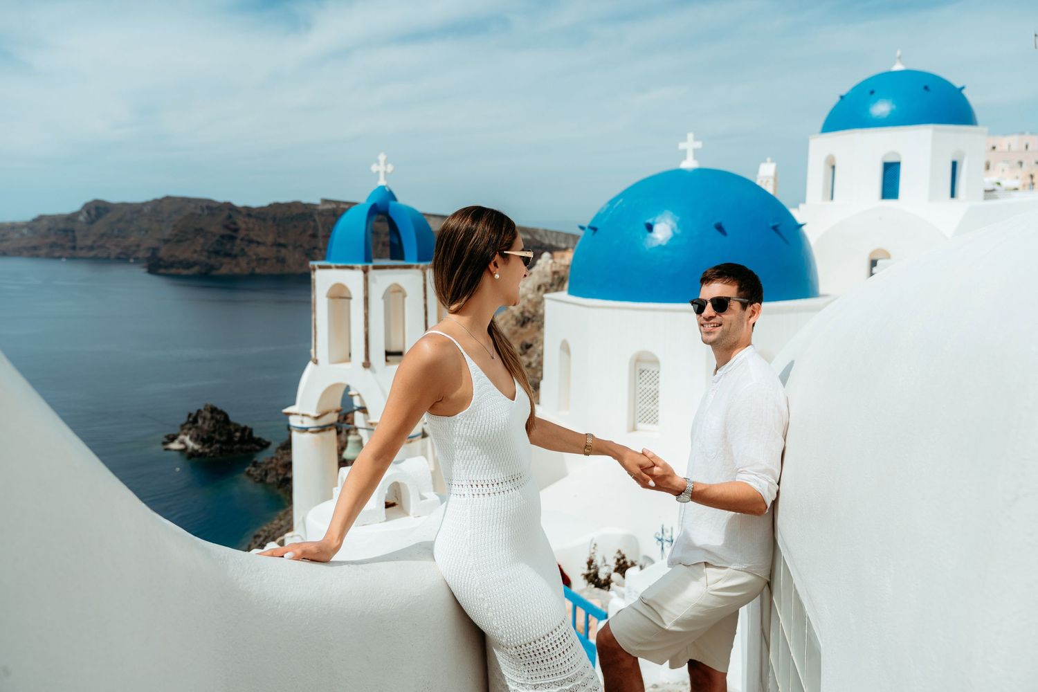 Couple holding hands on white terrace with iconic blue-domed churches in Santorini, Greece overlooking the Aegean Sea.