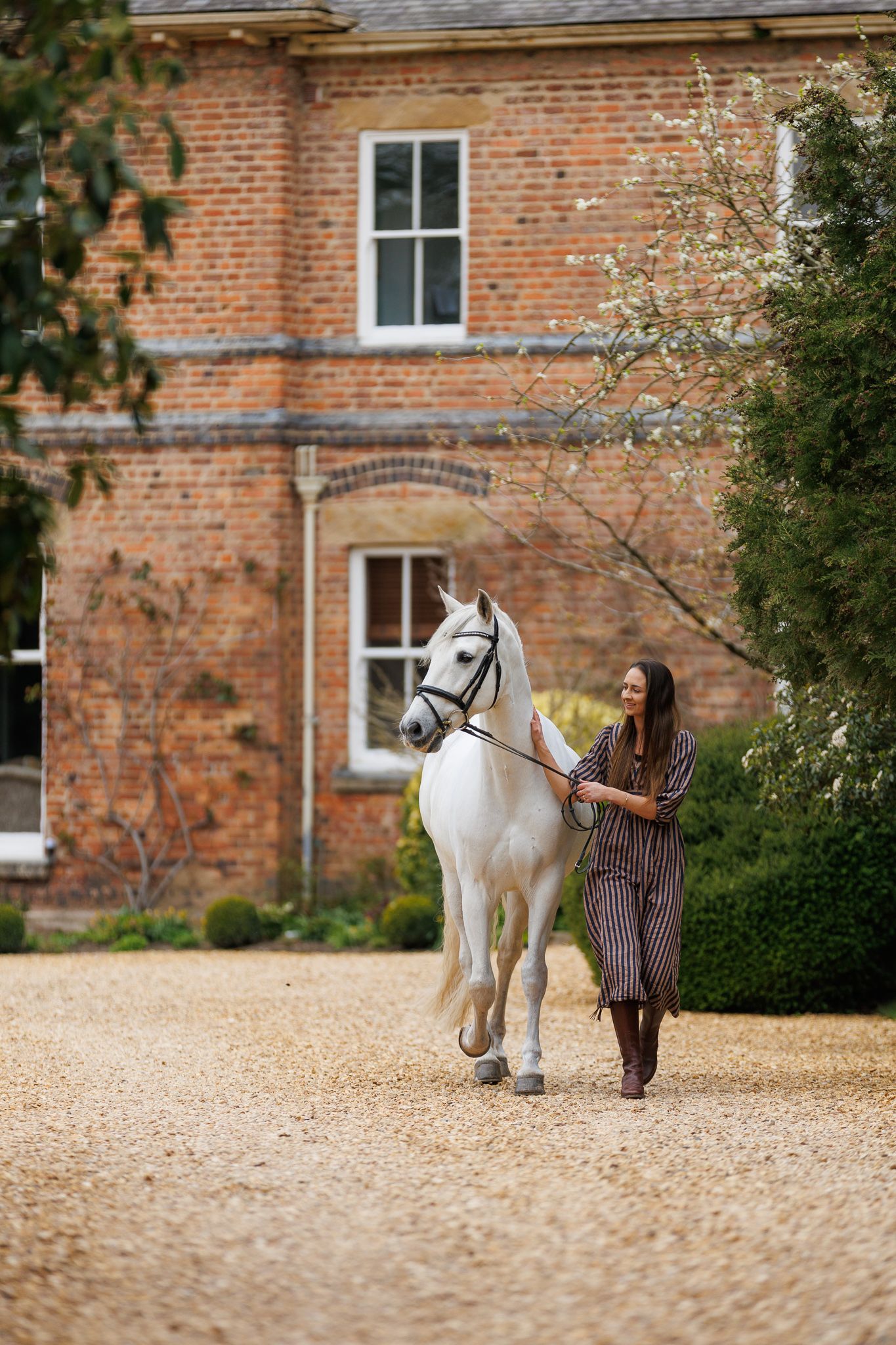 Young girl sitting on a stone wall between a chestnut horse and white pony, smiling outdoors.