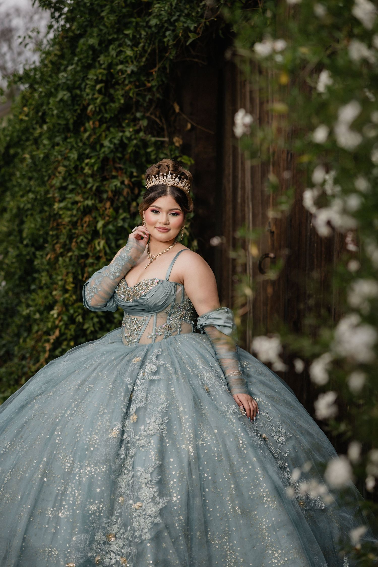 Young woman in a sparkling blue ball gown and gold crown poses gracefully outdoors among lush green ivy.