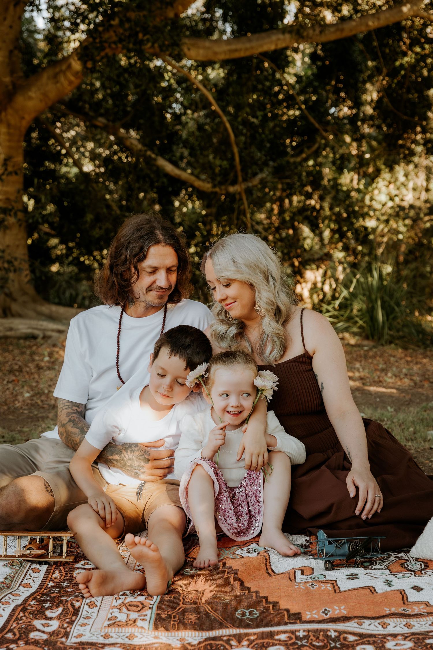 Family of four sitting on a patterned rug outdoors, parents smiling at two young children in a sunlit park setting.
