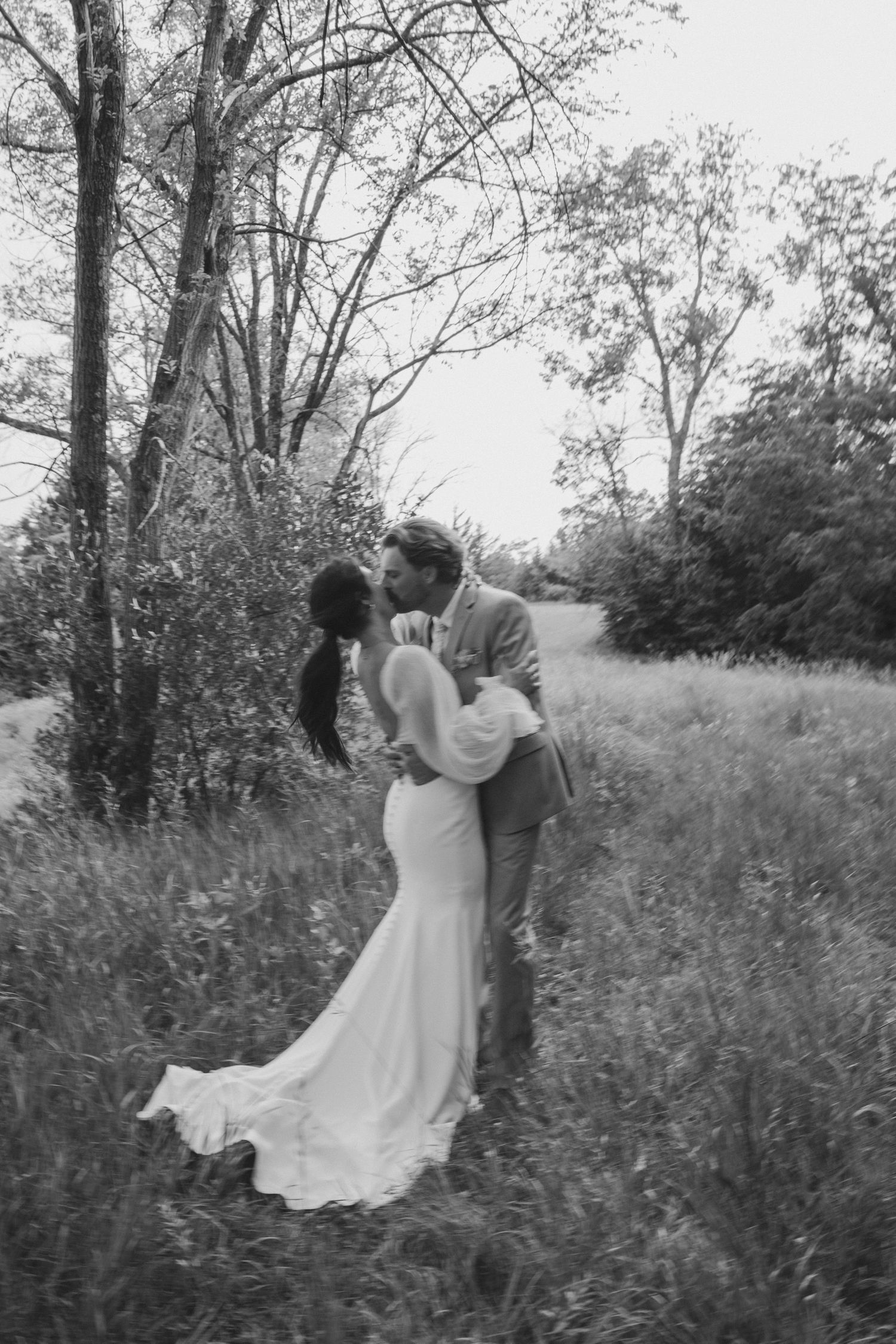 A romantic black and white photo series showing a couple embracing in a wildflower meadow surrounded by trees.
