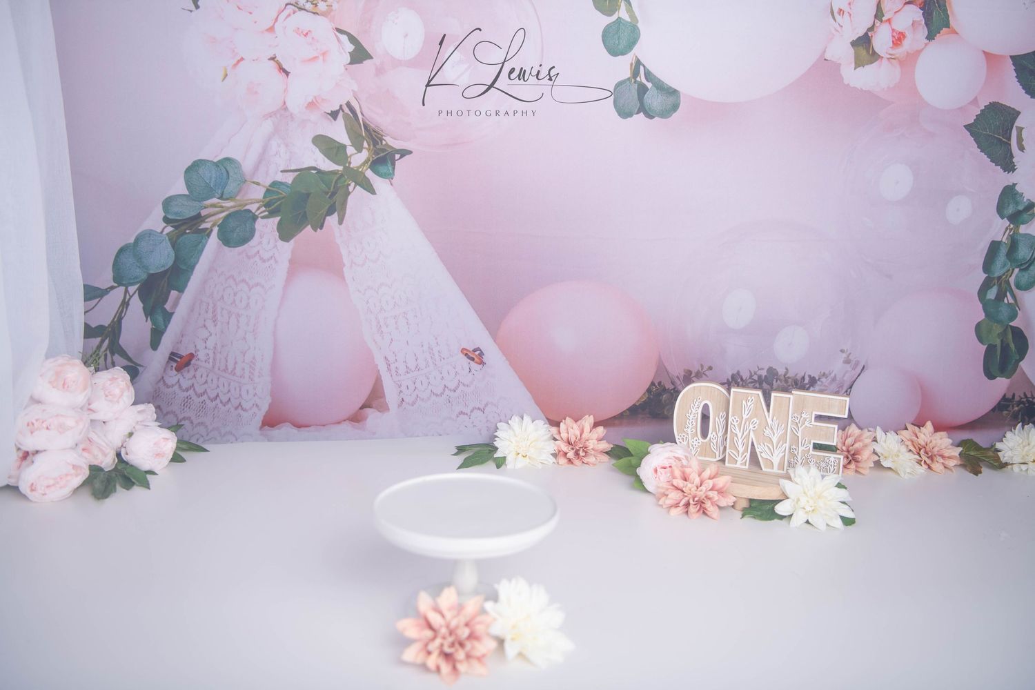 Pink first birthday cake smash setup with floral backdrop, balloons, white cake stand, and ONE letter sign.