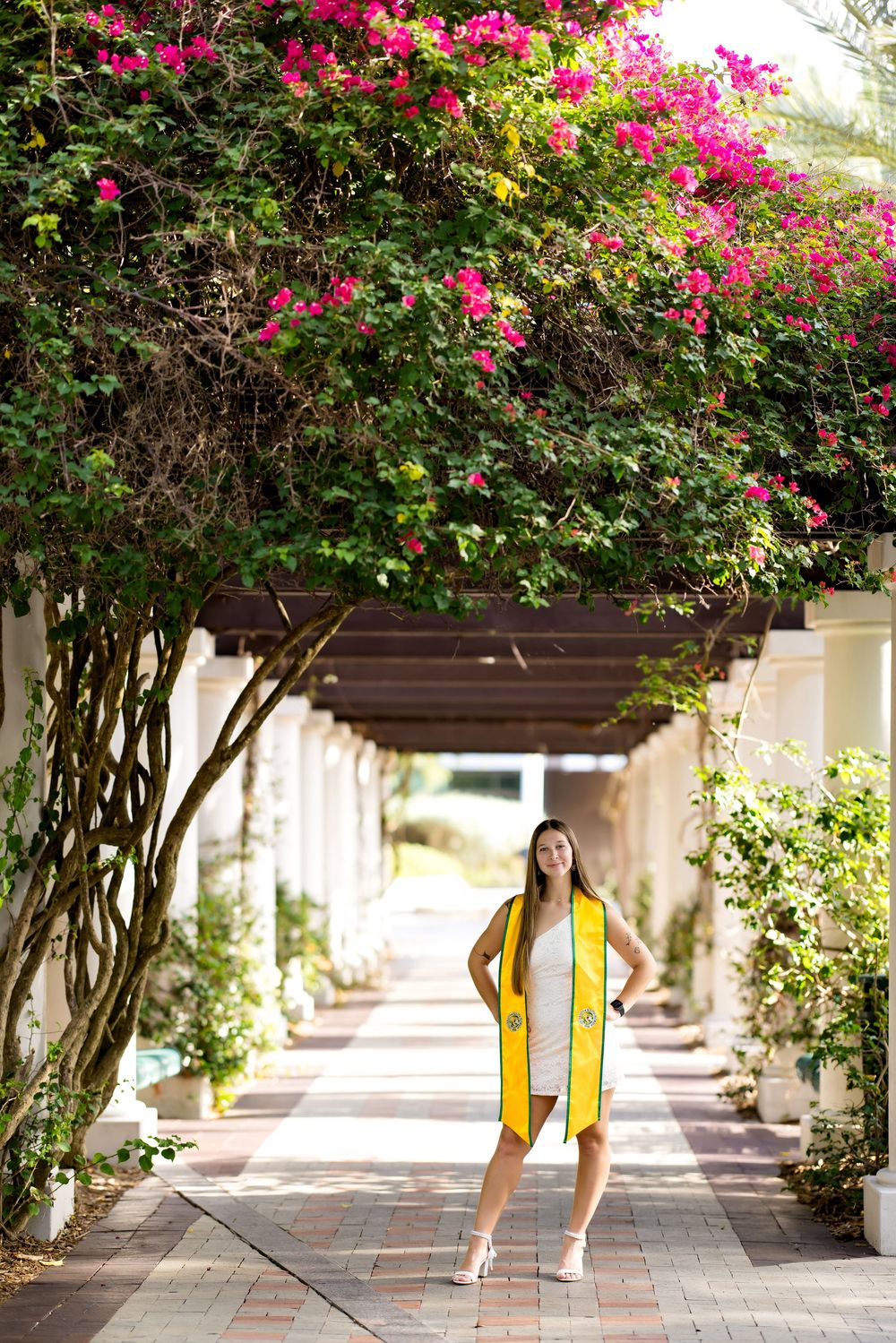 Graduate in yellow stole poses under flowering bougainvillea pergola on brick walkway.