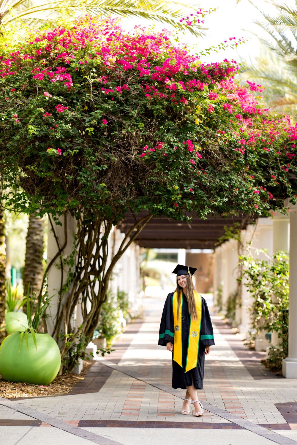 A graduate in black cap and gown with yellow stole walks under a bougainvillea-covered pergola on a sunny campus.