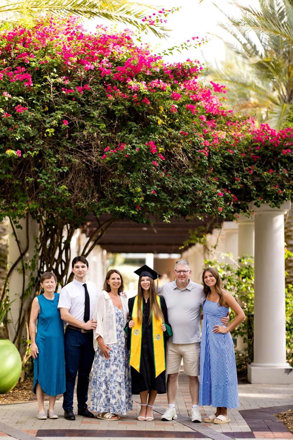 Family poses with graduate in cap and gown under blooming bougainvillea at outdoor graduation ceremony.