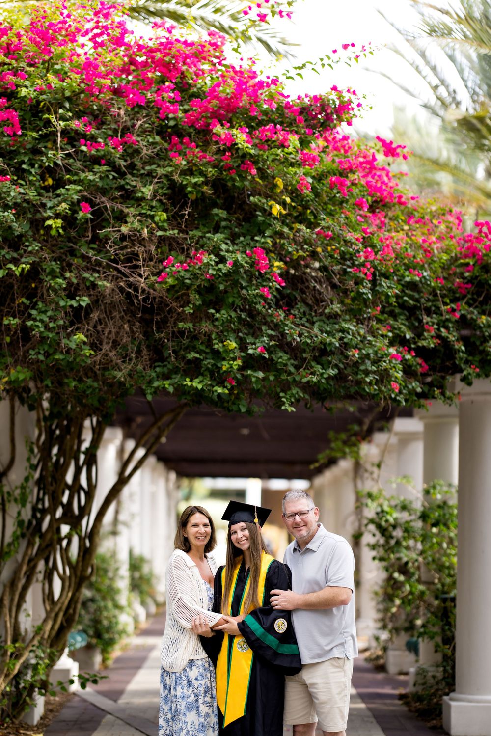 A graduate in cap and gown poses with two proud parents under a pergola adorned with vibrant pink bougainvillea flowers.