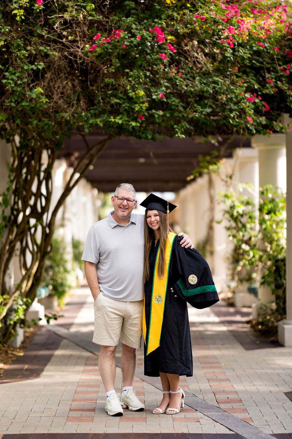 A proud graduate in cap and gown with yellow stole poses with an older man under a flowering pergola walkway.