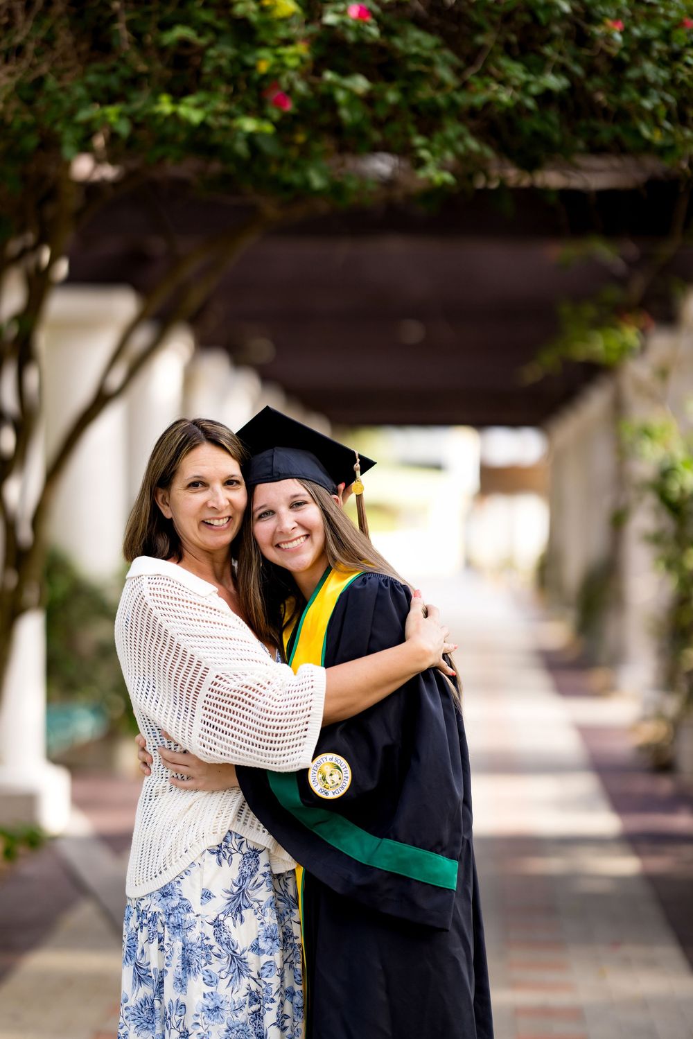 A proud graduate in cap and gown hugs a smiling woman in a floral skirt under a lush garden pergola.