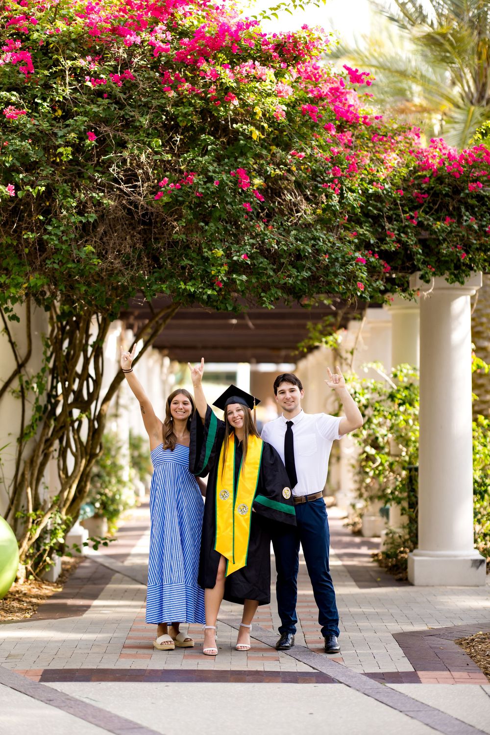 Three people celebrating a graduation outdoors under flowering bougainvillea, graduate wearing cap and green stole.