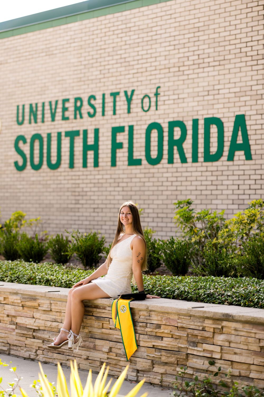 Young woman in white dress sitting on a stone wall in front of the University of South Florida sign.
