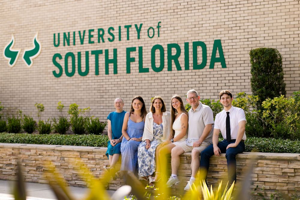 Group of six people posing in front of University of South Florida brick wall sign on campus.