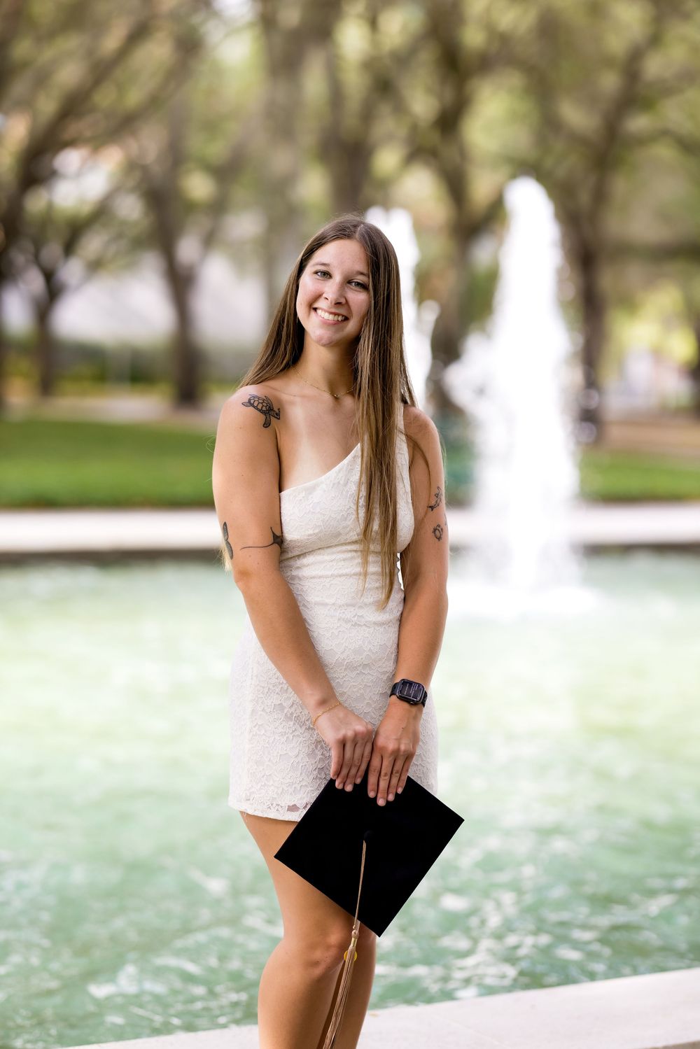 Young woman in white lace dress holding graduation cap, smiling in front of a fountain in a park.