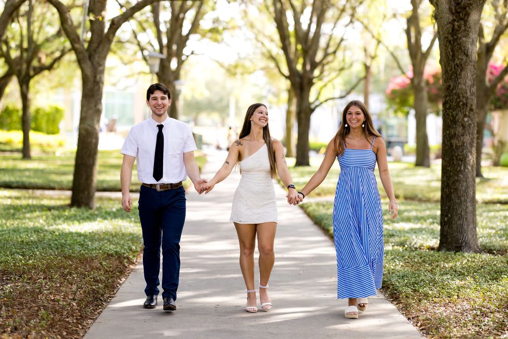 Three young adults walk hand-in-hand on a tree-lined path, dressed in formal attire for a special occasion.