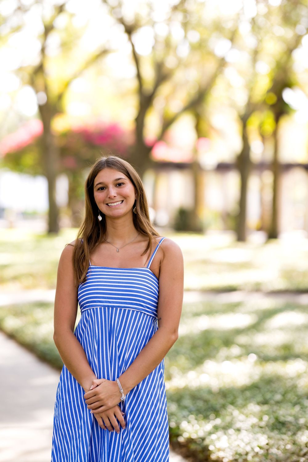 Young woman in blue striped sundress smiling outdoors in a sunny tree-lined park setting.