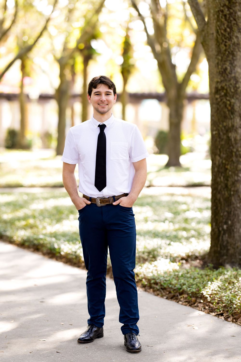Young man in white shirt and black tie smiling outdoors on a tree-lined pathway.