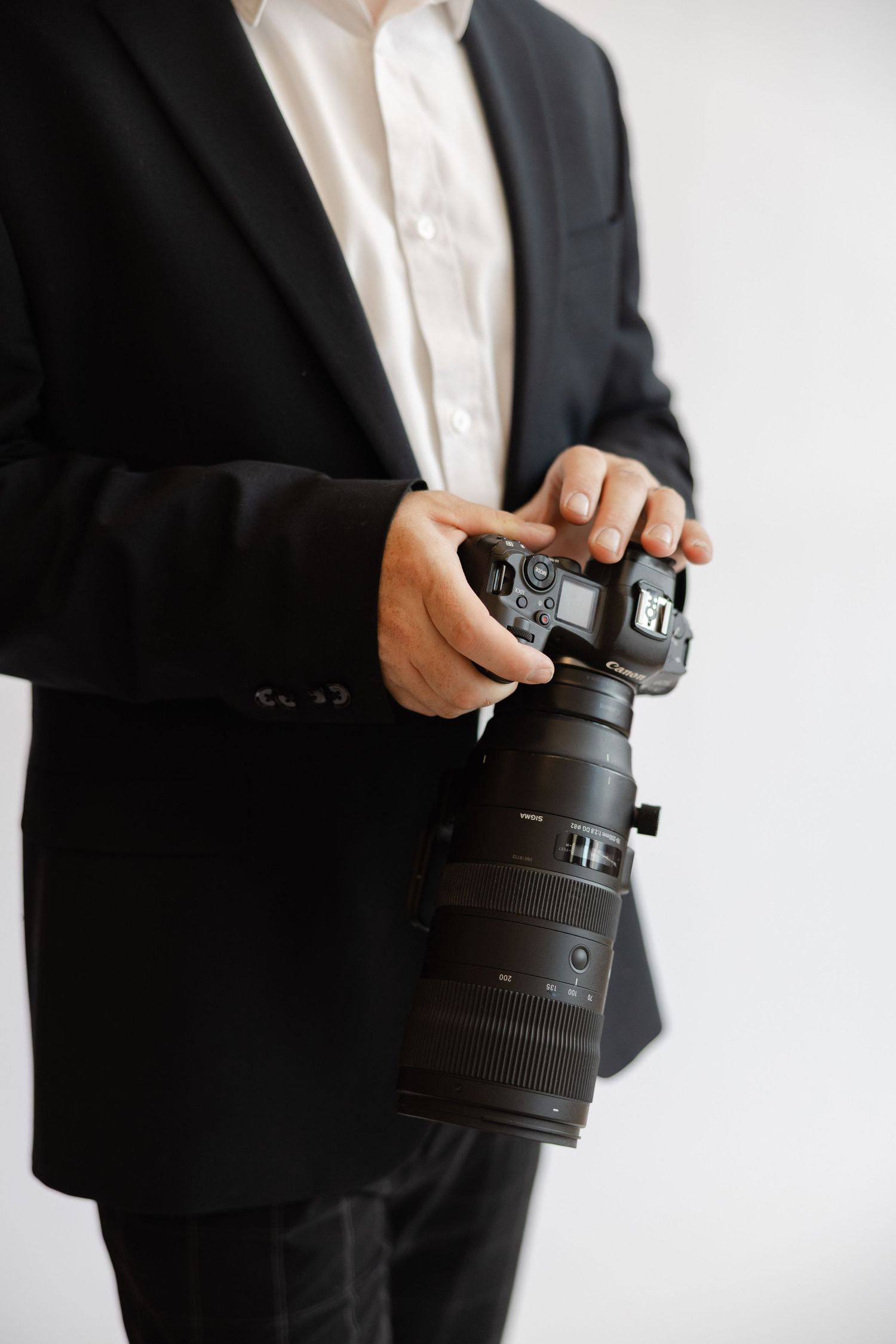 Professional photographer in black suit holding a DSLR camera with telephoto lens against white background.