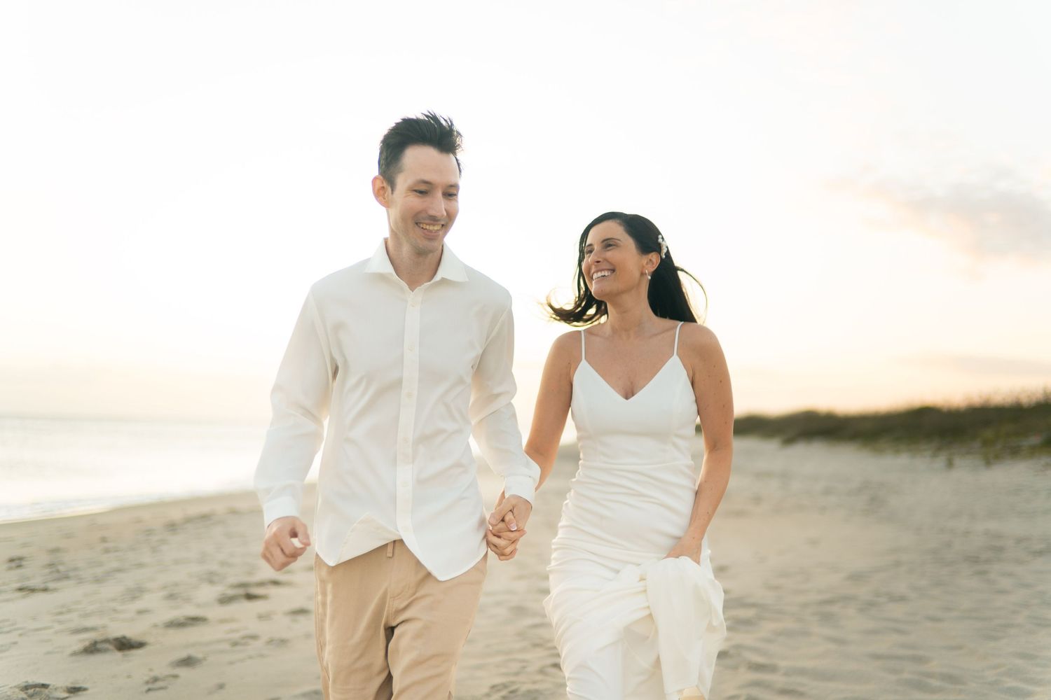 A couple in white attire walks hand in hand along a sandy beach at sunset, creating a romantic silhouette.