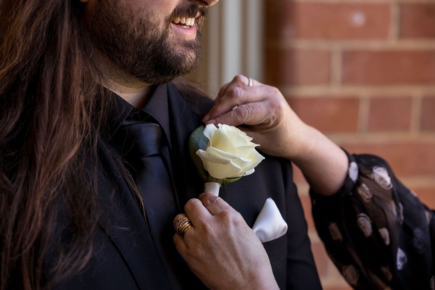 A close up image of a Mum's hands adjusting a white rose button hole flower on the Grooms black suit jacket.