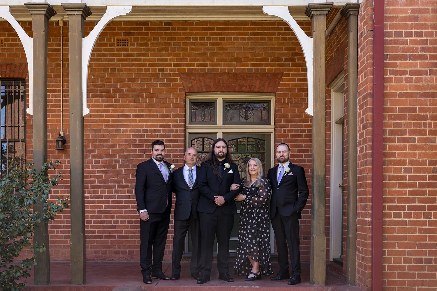 Group of wedding guests in formal attire gather outside a red brick building entrance.