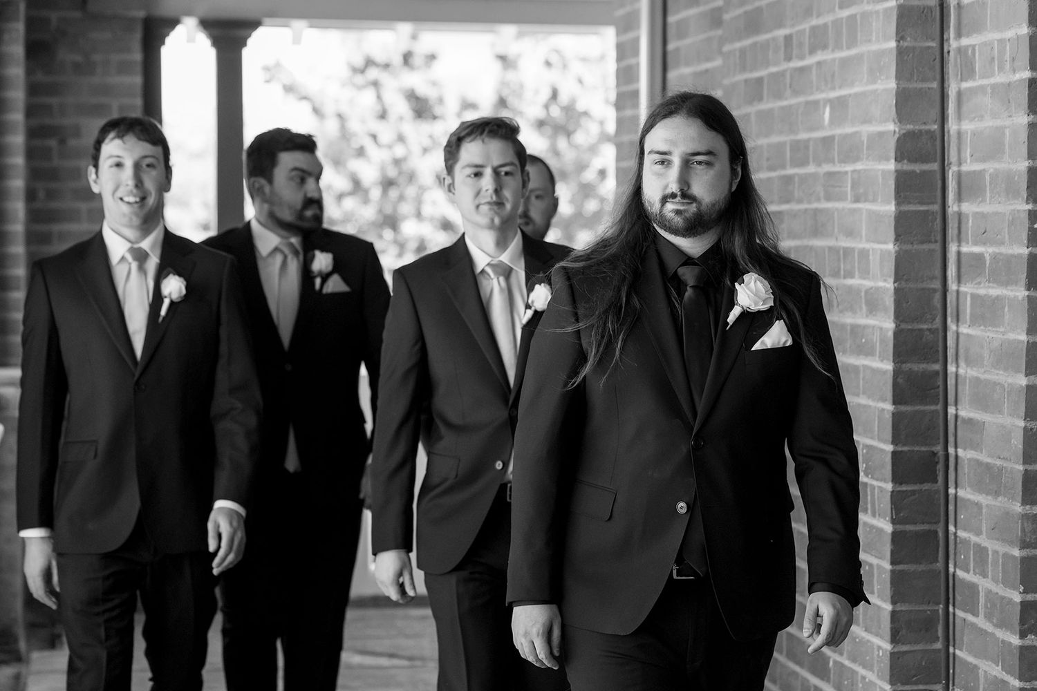 A group of groomsmen in black suits pose together along a brick wall for a formal wedding photo.