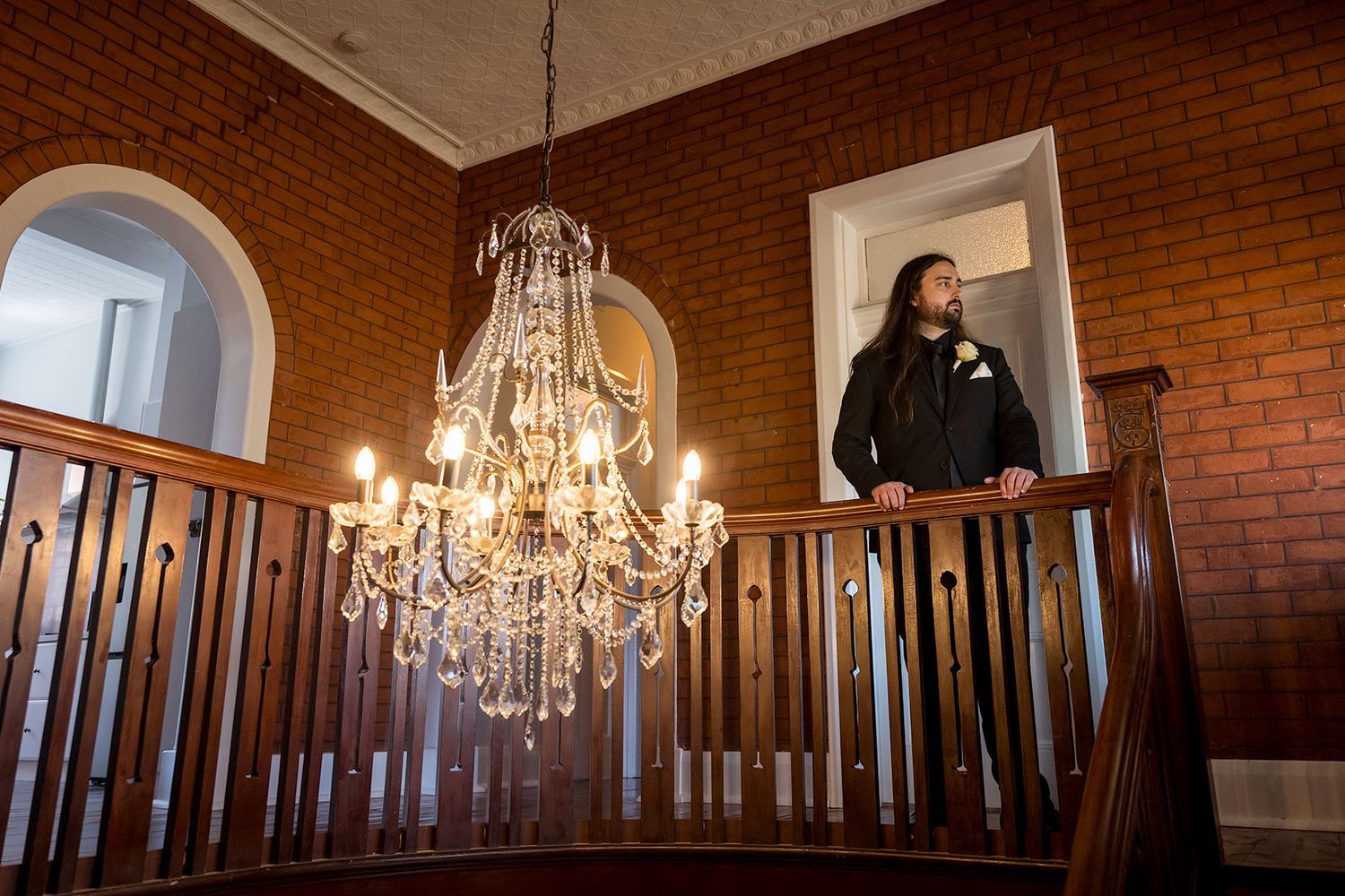 A Groom in black formal suit with a white rose button hole flower, behind an ornate railing with crystal chandeliers in the foreground.