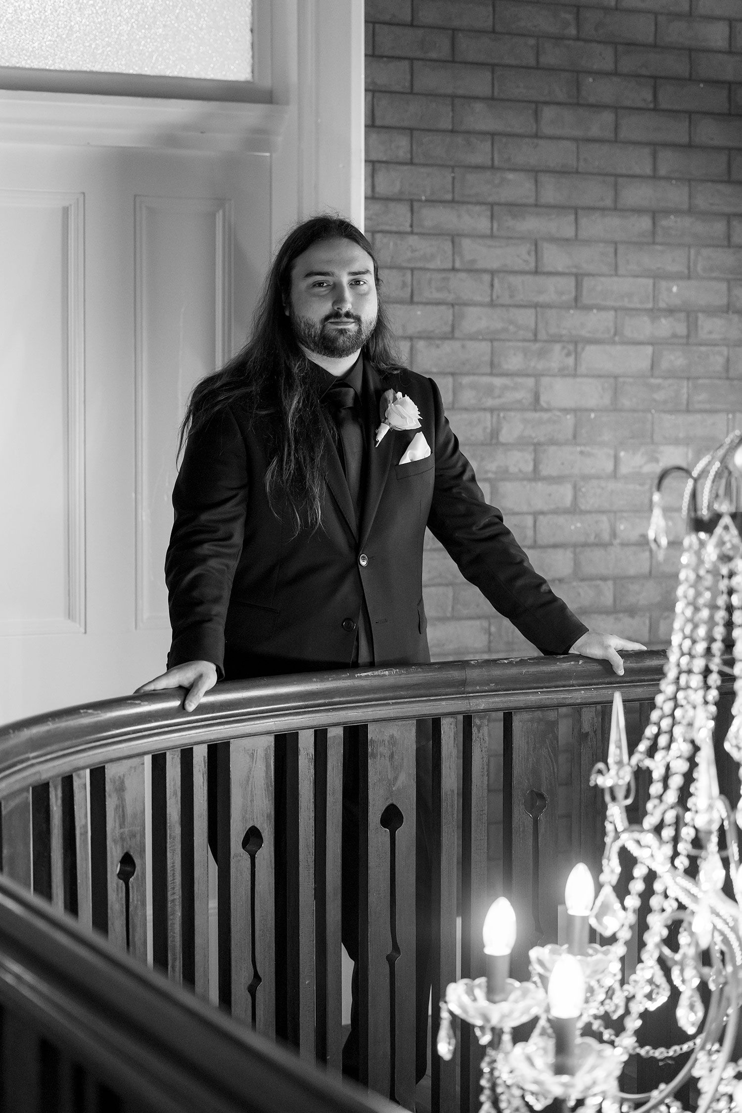 A Groom in black formal suit with a white rose button hole flower, behind an ornate railing with crystal chandeliers in the foreground.