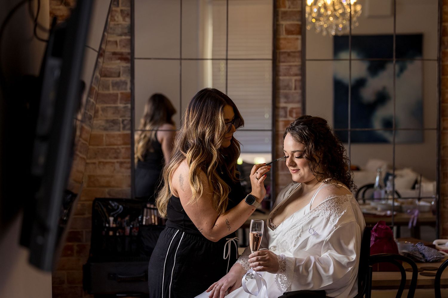 A bride during her make up session during wedding preparations in a white robe holding a glass of Champagne.