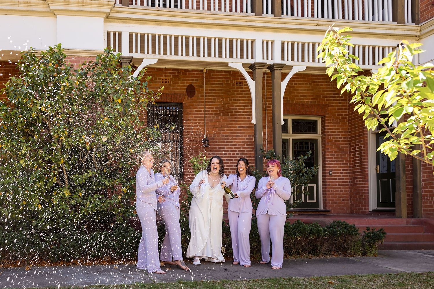 A bridal party in their PJs poses for photos in front of a brick building with arched walkways and greenery spraying a bottle of Champagne.
