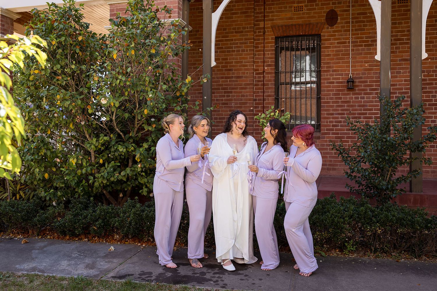 A bridal party in their PJs poses for photos in front of a brick building with arched walkways and greenery holding glasses of Champagne.