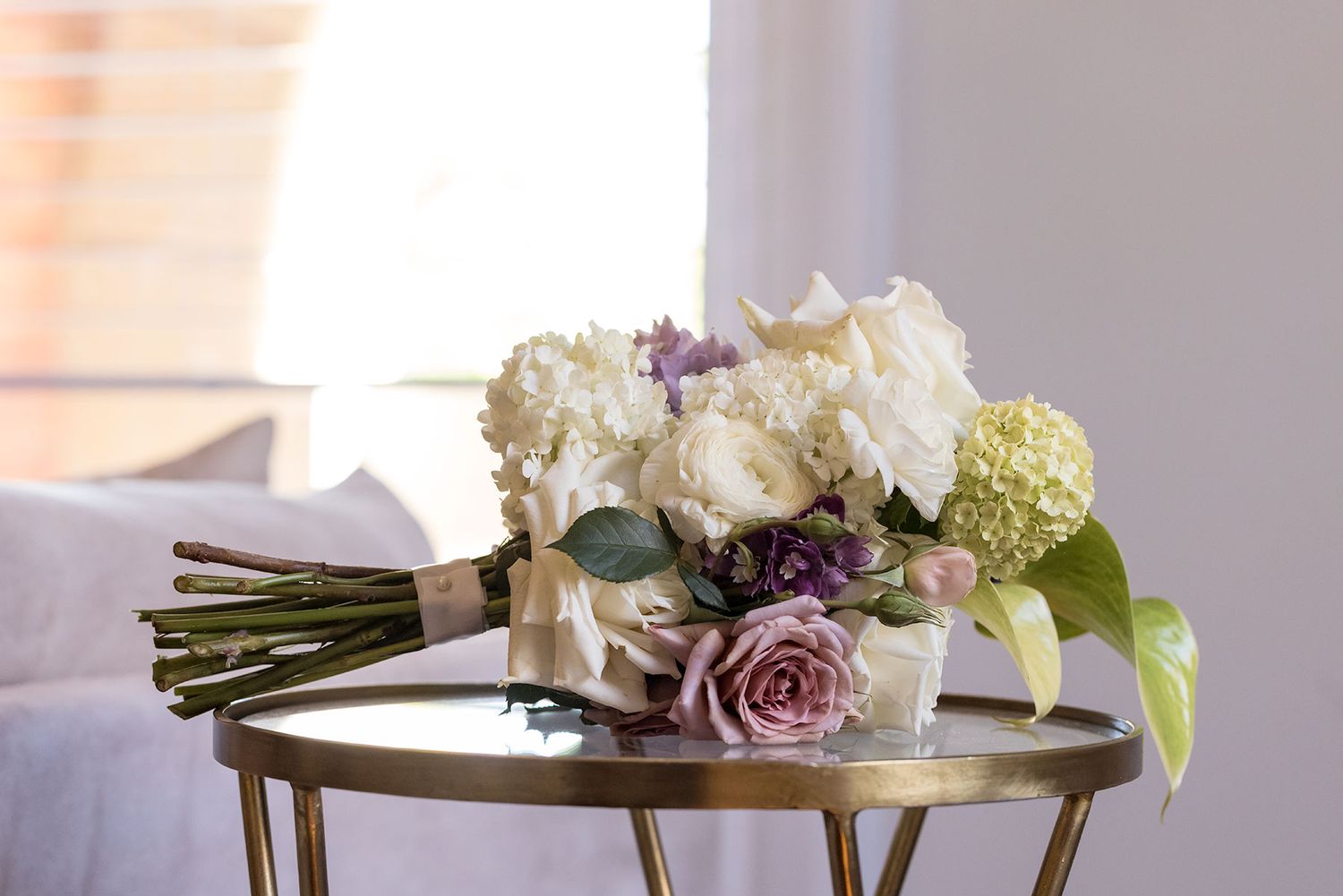 A delicate bouquet of white hydrangeas and dusty purple roses rests on a gold side table in soft natural light.