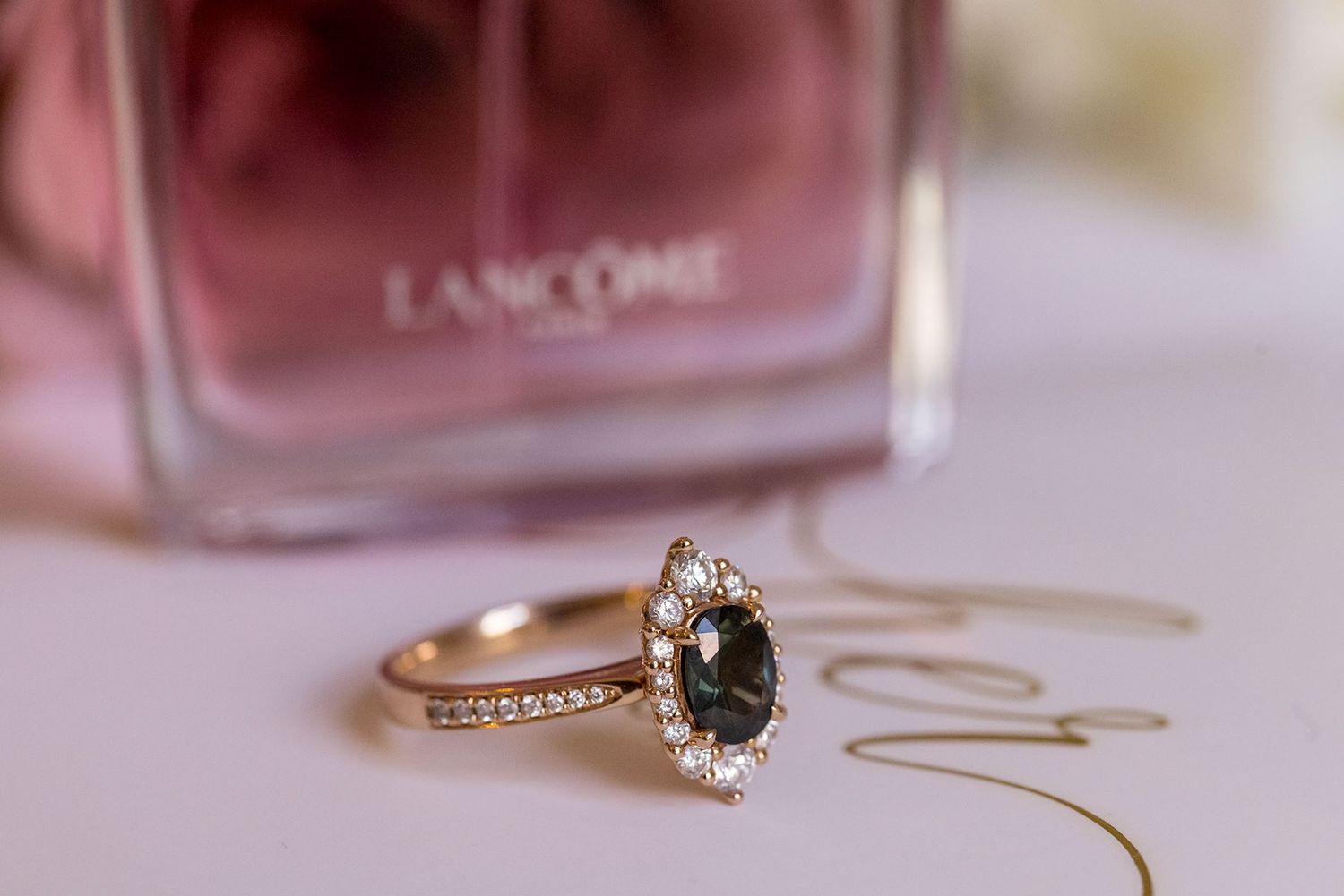 Gold engagement ring with dark center stone and diamond halo sits in front of pink Lancome perfume bottle.