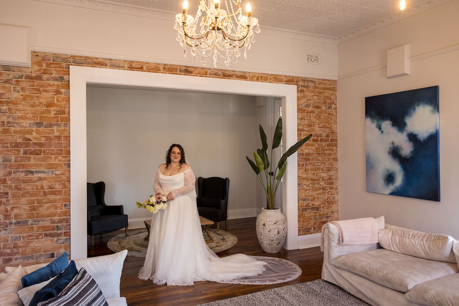 A portrait of a bride in an elegant wedding dress is displayed in a modern room with exposed brick walls, chandelier, and contemporary decor