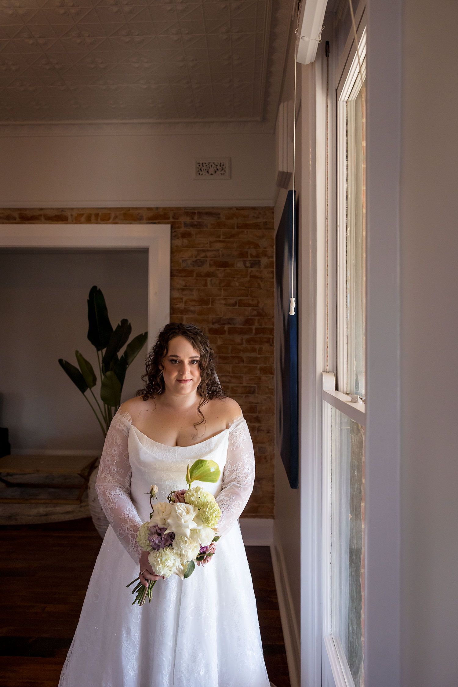A portrait of a Bride in her wedding dress holding a bouquet of flowers beside a window during her wedding preparations.