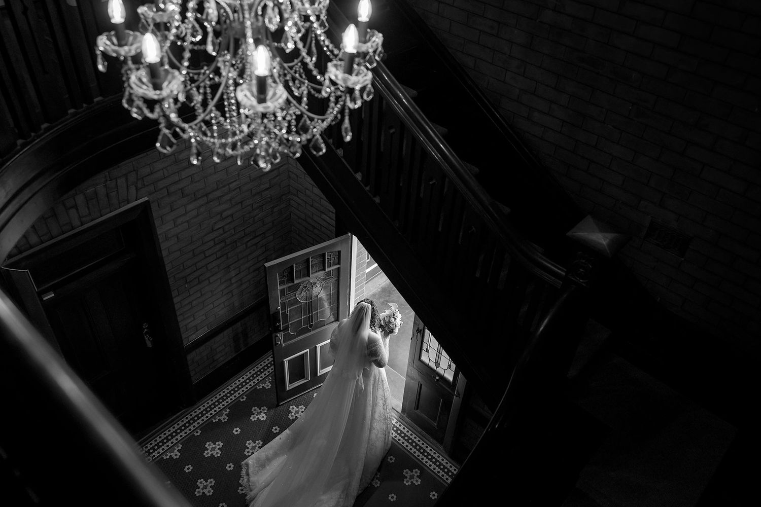 An elegant crystal chandelier hangs above a black and white wedding scene photographed from above with a bride walking out in her gown.