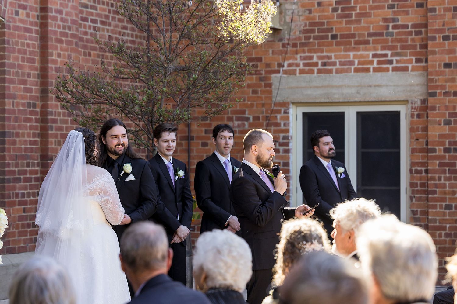 A wedding ceremony takes place outdoors against a red brick building on a sunny afternoon at the Corowa Distilling Co.