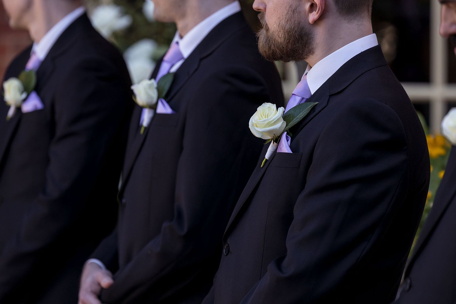 Groomsmen in black suits with purple ties and white boutonnieres stand in line at a formal wedding ceremony.