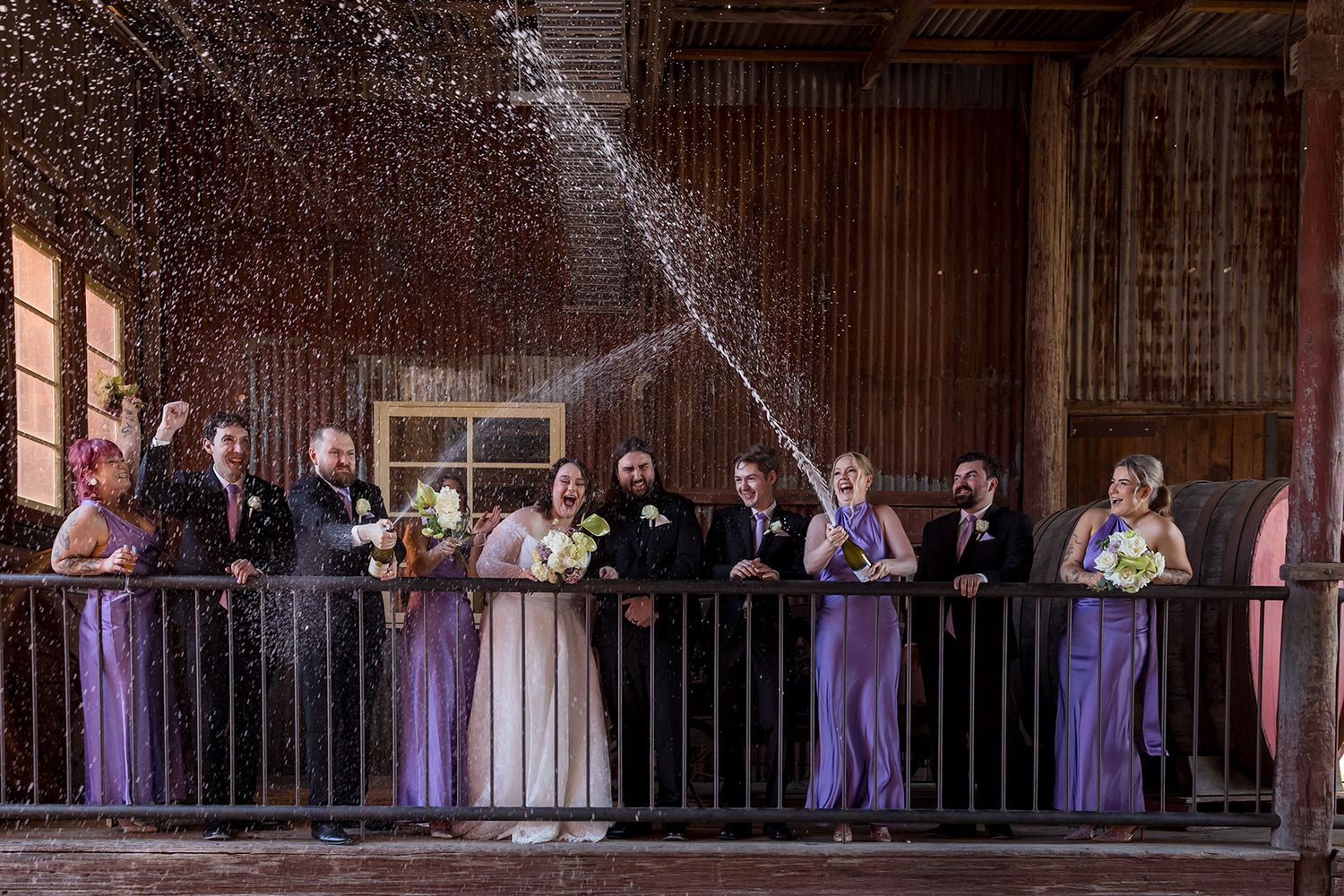 Wedding party members in purple dresses stand along a rustic wooden balcony during a celebration spraying bottles of Champagne.