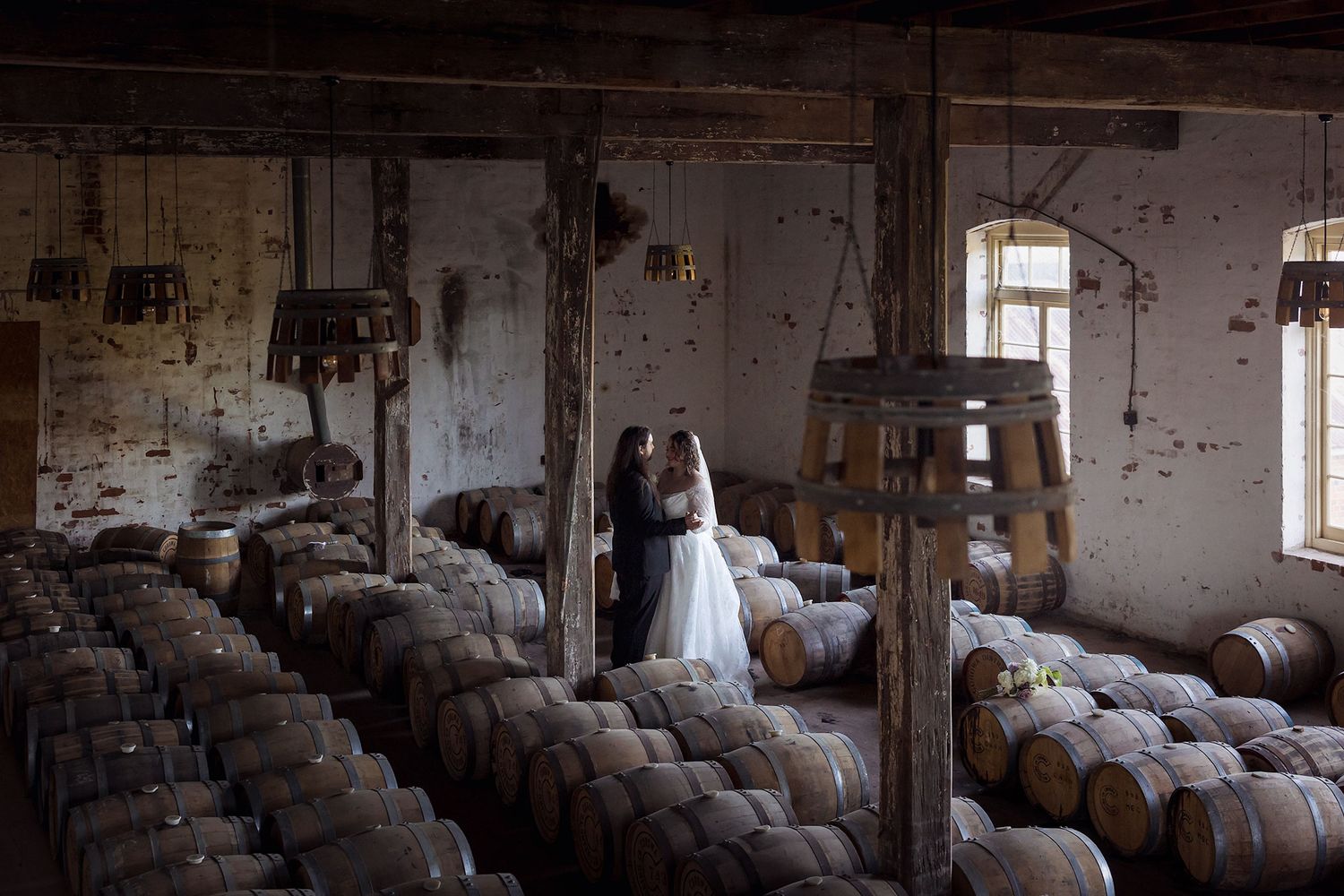 A Bride and Groom in rows of wooden whiskey barrels age in a dimly lit distillery warehouse with sunlight streaming through windows.
