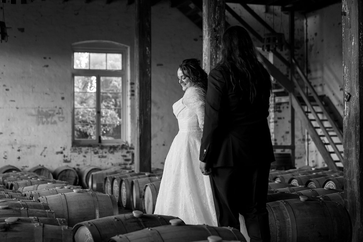 A Bride and Groom in rows of wooden whiskey barrels age in a dimly lit distillery warehouse with sunlight streaming through windows.